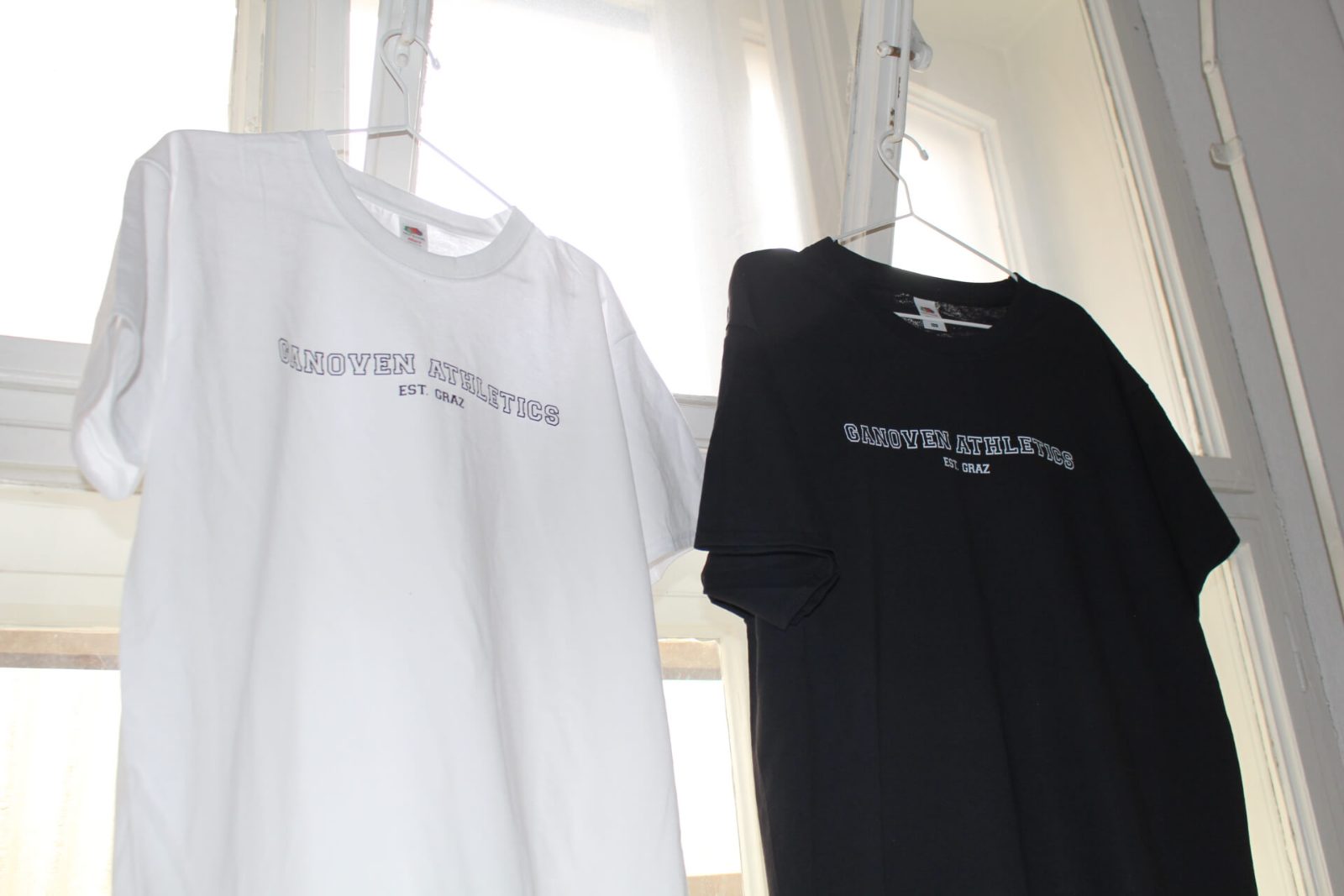 Weißes und schwarzes Merchandise-T-Shirt mit „Ganoven Athletics Est. Graz“-Siebdruck auf Kleiderbügeln am Fenster